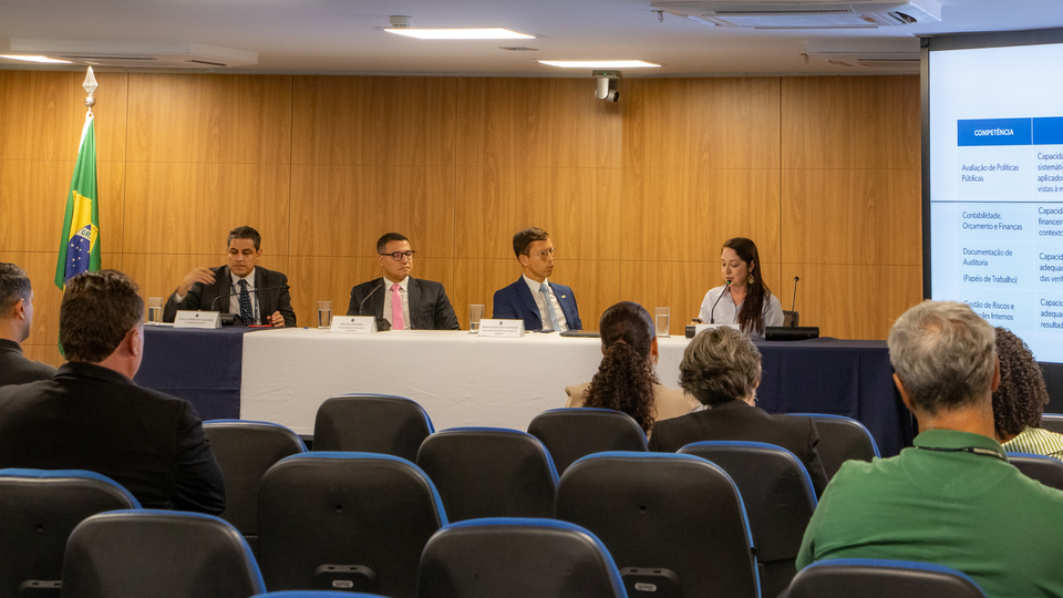 Três homens de terno e uma mulher de roupa clara compoem mesa dentro de um auditório